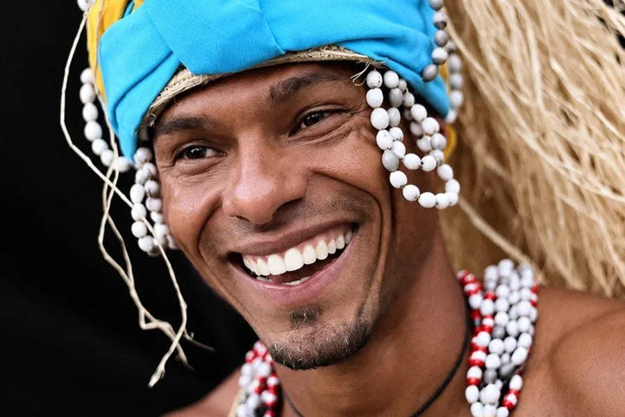 Mestre Letho Nascimento souriant, avec sa coiffe turquoise et ses colliers de perles